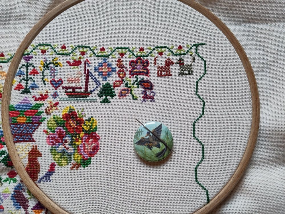 Photo of a cross stitch in progress in a wooden hoop. It's a colorful sampler with a strawberry border full of traditional and funky motifs, like two small dogs, a flower bouquet and a really funky flower.