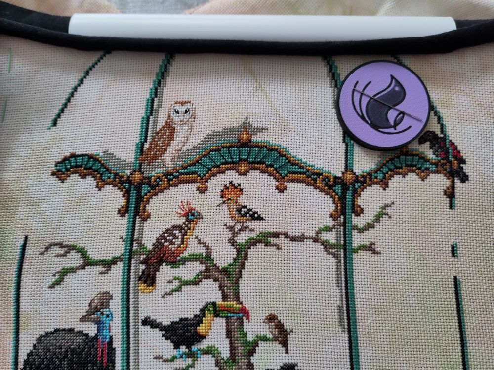 Photo of a cross stitch in progress. A couple of colorful birds in a huge ornate aviary.