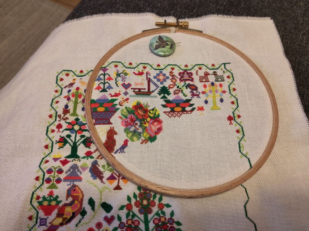 Photo of a cross stitch in progress in a wooden hoop. It's a colorful sampler with a strawberry border full of traditional and funky motifs, like two small dogs, a flower. The funky tree is completed with funky oblong fruits and another berry bowl is started