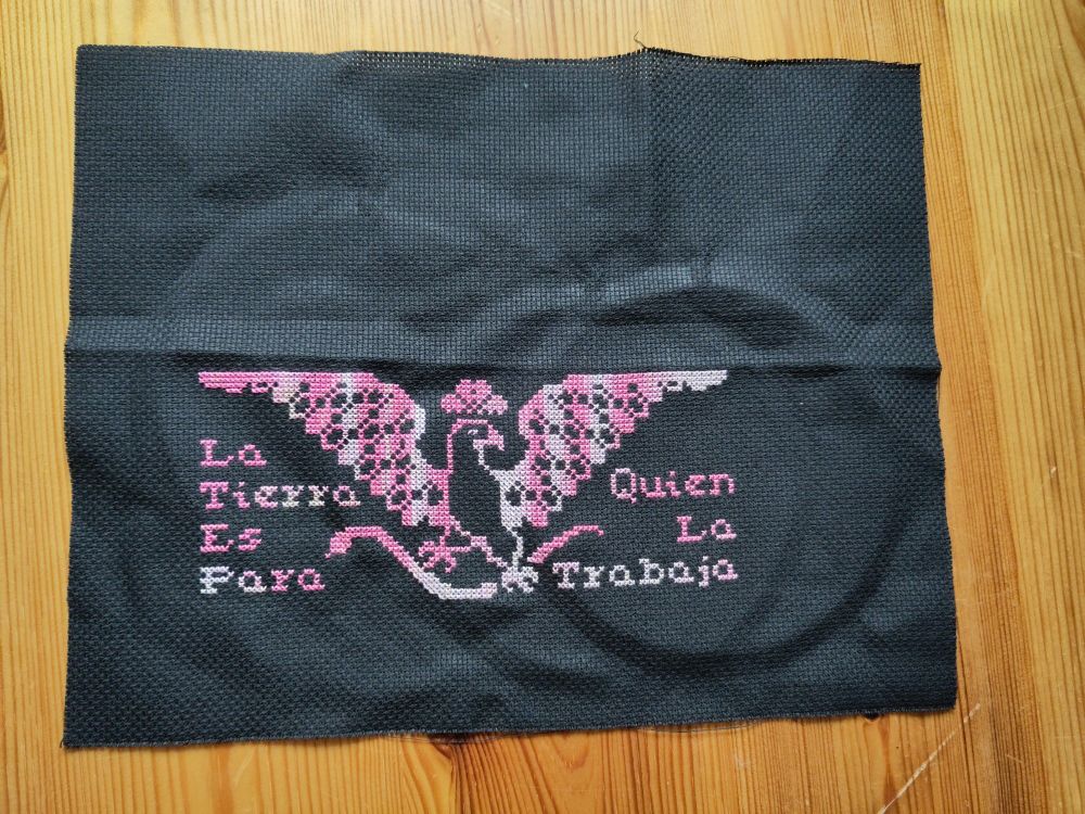 Photo of a cross stitch with a variegated pink to white thread on black fabric. A big bird with wings wide open is holding a snake in its fangs. On the sides of the bird are the words "La Tierra Es Para Quien La Trabaja".