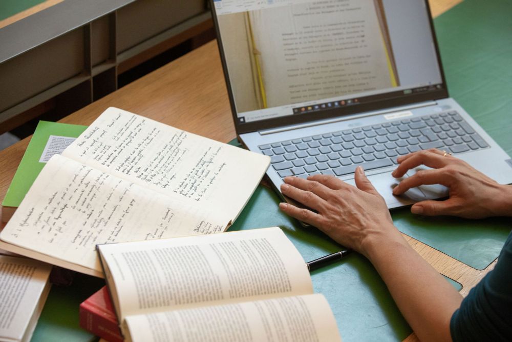 Une personne travaille dans la bibliothèque de l'IHA à partir de notes manuscrites, de divers ouvrages et de son ordinateur portable.