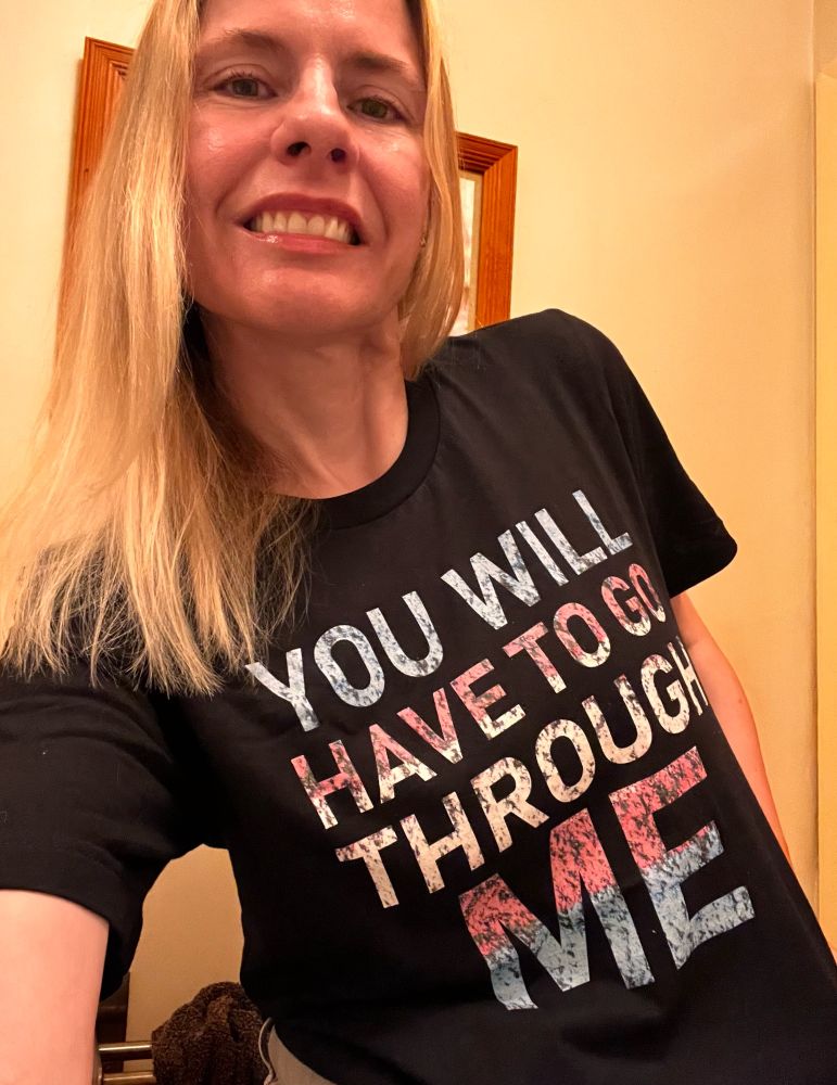 Me in a black T shirt, the front print reads YOU WILL HAVE TO GO THROUGH ME in trans pride colours