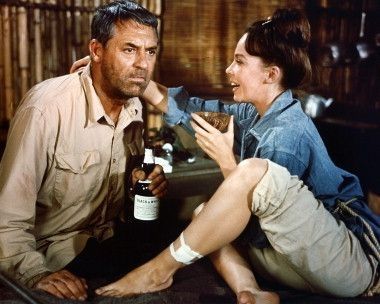 Fotograma de la película "Father goose" en la que aparecen los protagonistas, Cary Grant y Leslie Caron, los dos sentados uno enfrente del otro, él con una botella en la mano y ella sujetando un coco a modo de vaso, y con un vendaje en el pie.