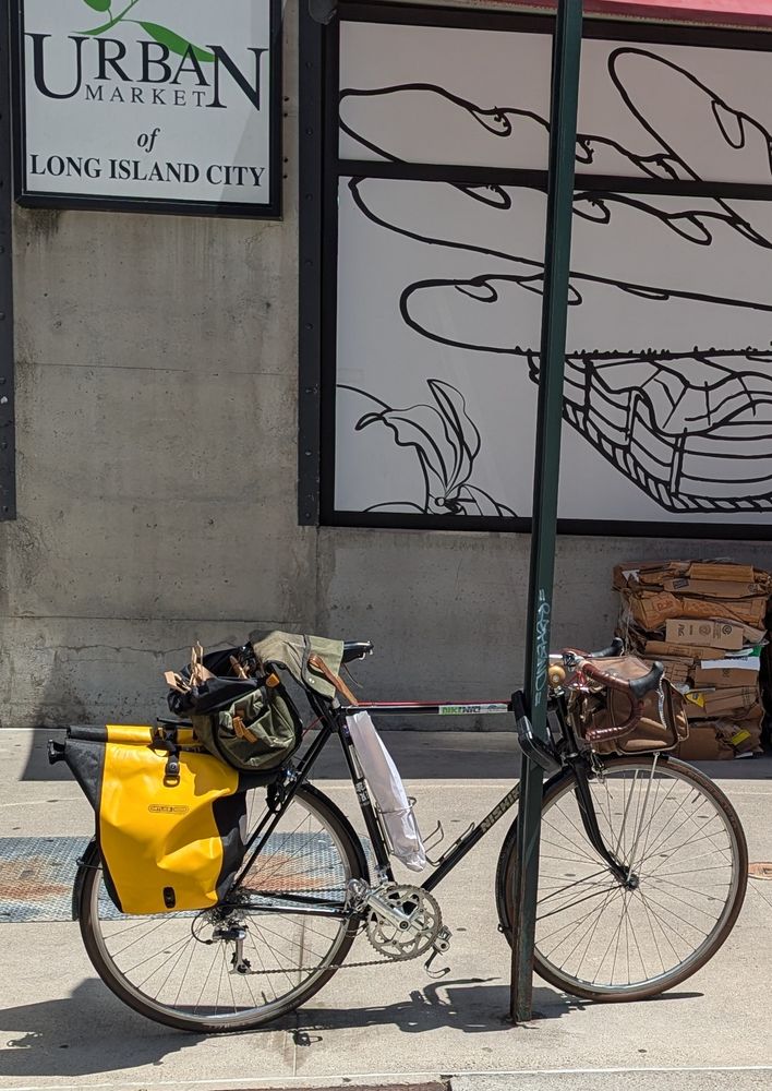 A black bicycle with all manner of luggage (front bag, giant saddle bag, and panniers) is locked to a street sign in front of a supermarket. A baguette is in the bikeçs bottle cage.