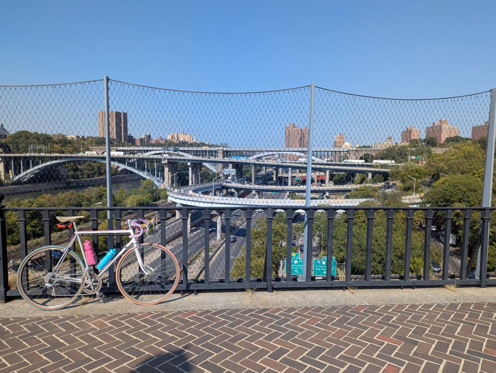 A silver and white bicycle leaned up against a fence. Three or four highways are visible in the background, overlapping and intertwined.