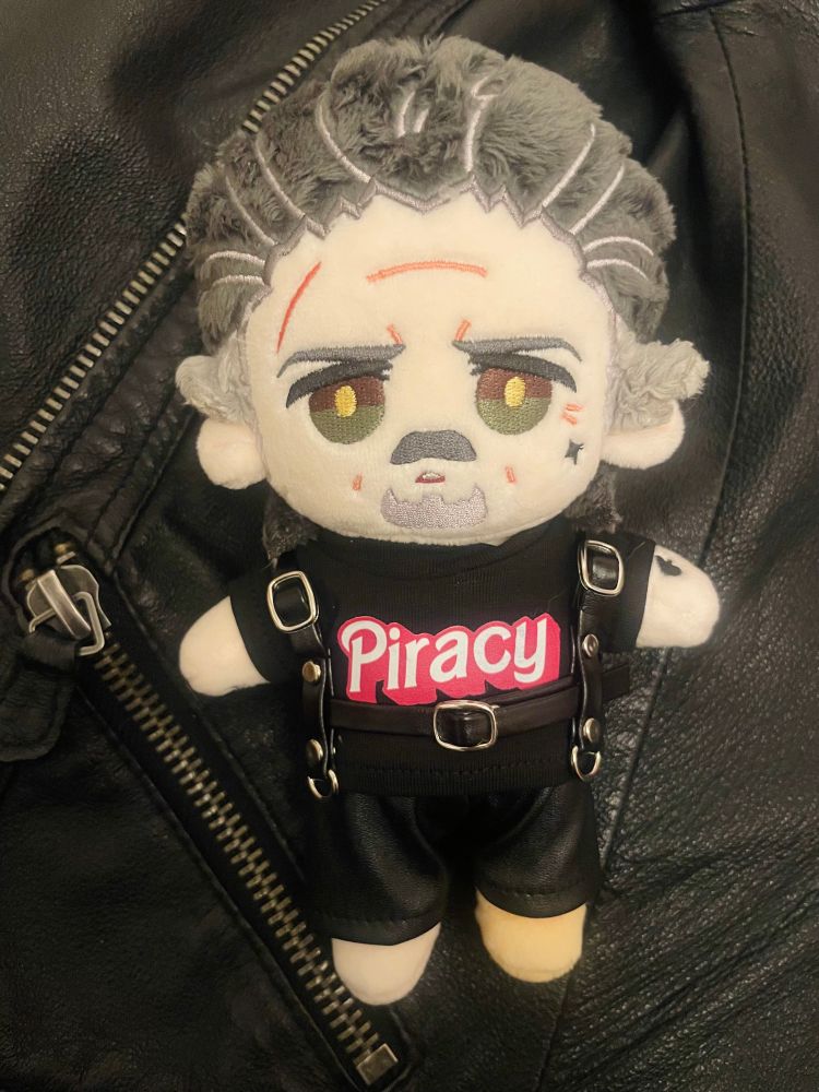 Izzy doll by Rights on a background of black leather. He’s surrounded wears leather trousers, a leather harness, and a black T-shirt emblazoned with “piracy” in the pink Barbie font