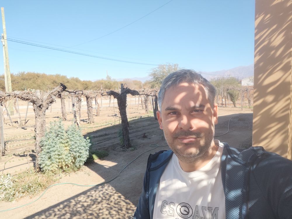 Selfie of me in front of a backyard garden with vines, with no leaves or grapes as it is winter. In the background some mountains can be seen. Cafayate, Salta, Argentina.