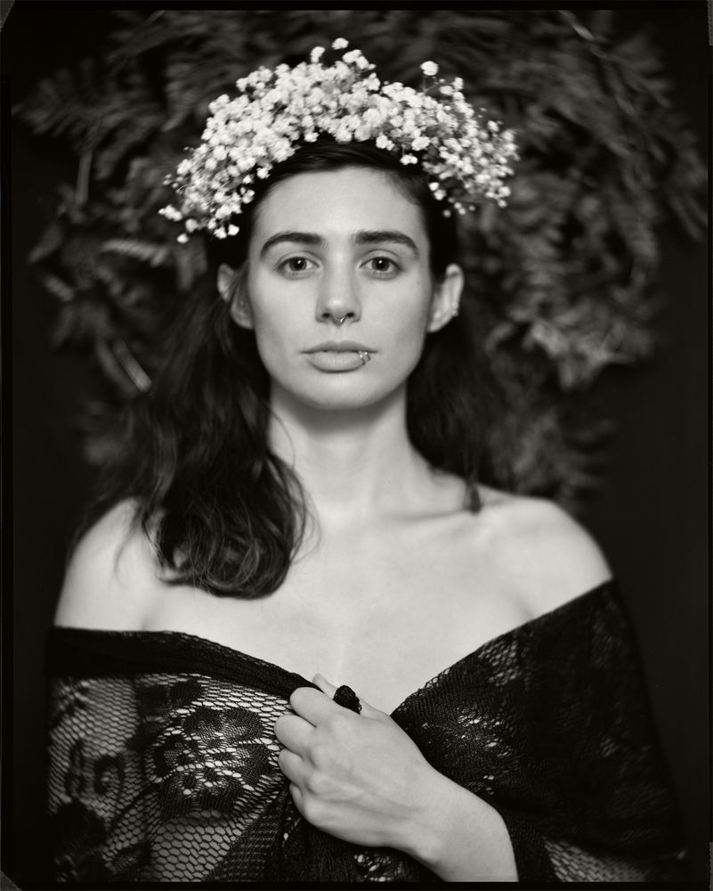 A black and white photographic portrait of a woman wearing a black lace shawl and a flower crown.