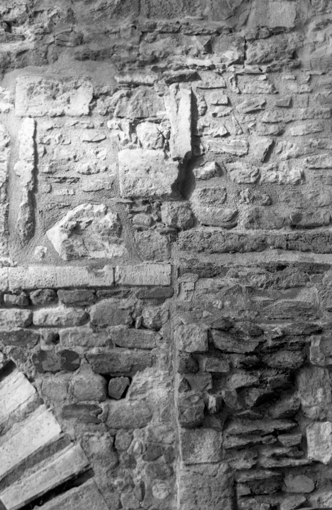 Black and white photograph of a stone wall, the edge of an arch in the lower left corner and much evidence of construction at different times