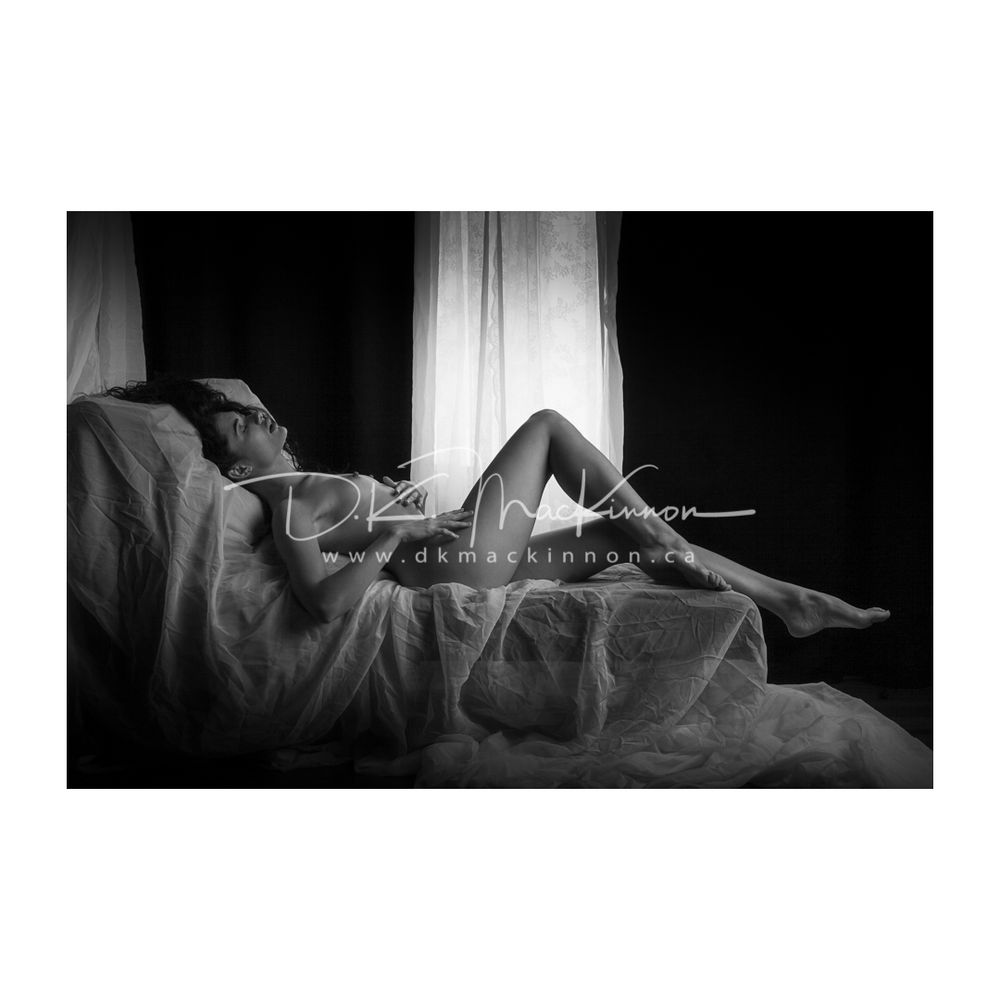 Black and white image of a nude woman with long curly hair lying on her back on a bed in front go a narrow window with patterned shear drapes. Her head is tilted back and the leg closest to the viewer is raised into a triangle.