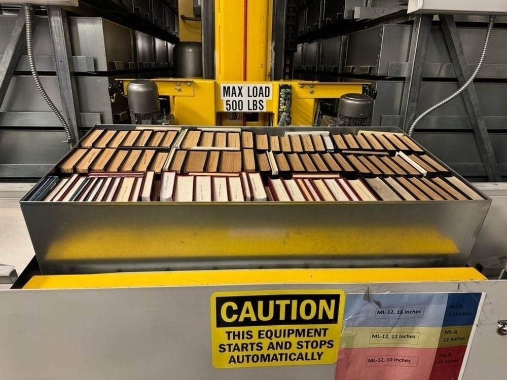 A metal tray of roughly 100 books sits on a giant yellow arm of a machine that transports the books. Other metal trays of books are in the background. A caution sign notes that the equipment starts and stops automatically.