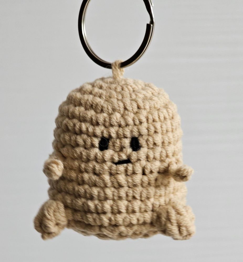 짧은 팔다리가 달린 감자 모양의 뜨개인형. 키링에 매달려 있다.