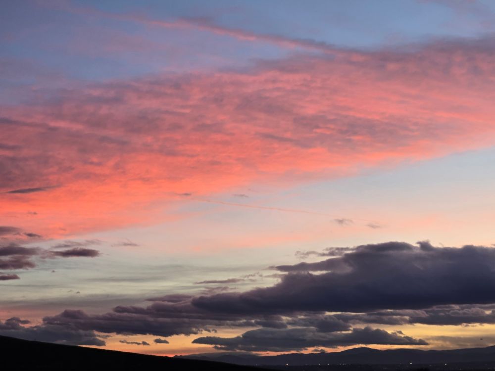 Pink clouds over dark grey clouds in front of the end of an orange sunset