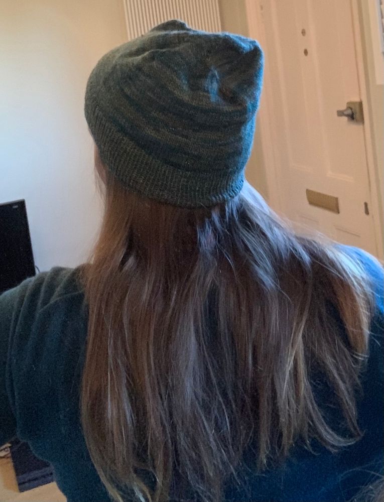 Mirror selfie of the back of a woman with long brown hair wearing a hand knit slouchy beanie in a green, teal, gold variegated yarn. 