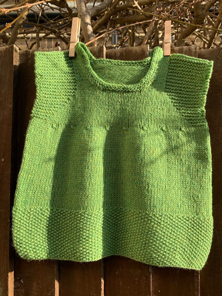 A pea green hand knit baby vest with garter stitch detailing at the shoulders and a long moss stitch hem. It is pinned with clothespins to a brown wooden fence in the sun. 