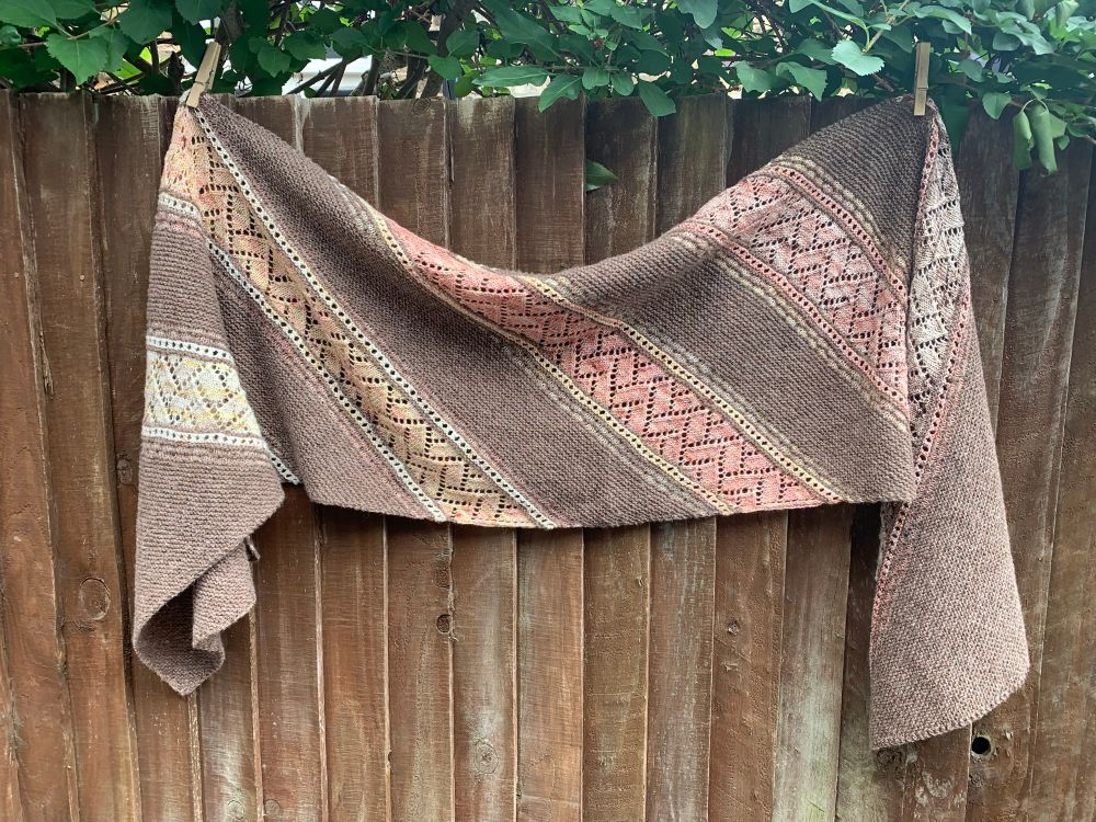 A rectangular hand knit shawl pinned to a brown wooden fence. It is mostly brown with stripes in lace in a gradient from light yellow through coral to brownish coral. 