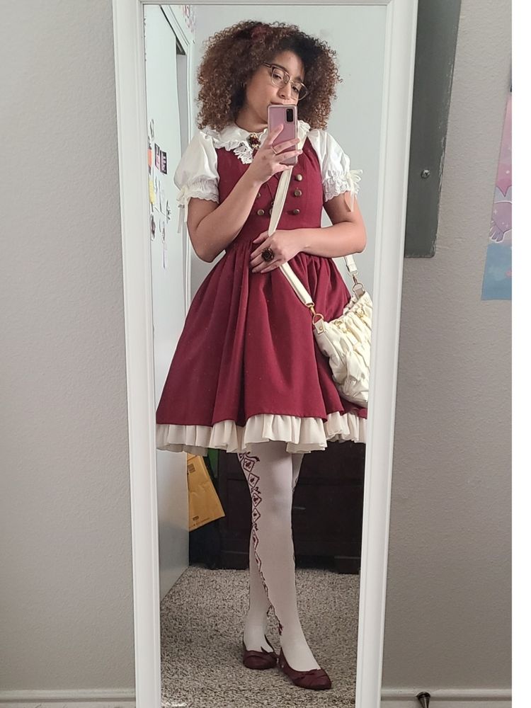 Dany poses in front of a mirror. Shes wearing a red JSK with a white blouse. The JSK has brass colored buttons on the bodice and has a tan ruffle on the hem. Shes wearing OTKs with a diamond and rose motif that goes up the side and red flats. She also has a cream cross body purse.