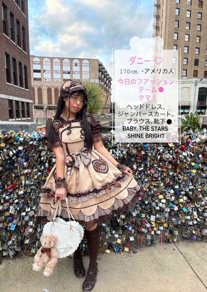 Dany wears a tan colored JSK that has bear ears on the straps. It also has a mix of patterns like gingham and polkadots. She has a matching headdress, a brown blouse, brown socks and brown flats. She has a white, heart scalloped purse with a tan teddy bear and a brown bow. There's a text: ダニー、170cm、アメリカ人、今日のファッションチム:クマ、ヘッドドレス、ジャンパースカート、ブラウス、靴下: baby the stars shine bright. She's standing in front of a fence that is covered in locks. 
