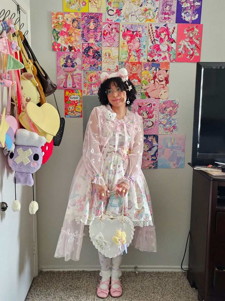 Dany poses by holding her purse in front of her with both hands. She is wearing a white bear headdress with pink bows, a pink peignoir, a white jsk with pastel prints of bears, rocking horses, stars, bunnies and candy, white tights with similar pastel motifs, white crew socks topped with lace and pink flats. She also has a white, scalloped heart bag with a star charm, a bear brooch and pastel bracelets and rings. Her background is filled with saturated posters mostly of Tokyo Mew Mew and she has a myriad of bags to her side. 