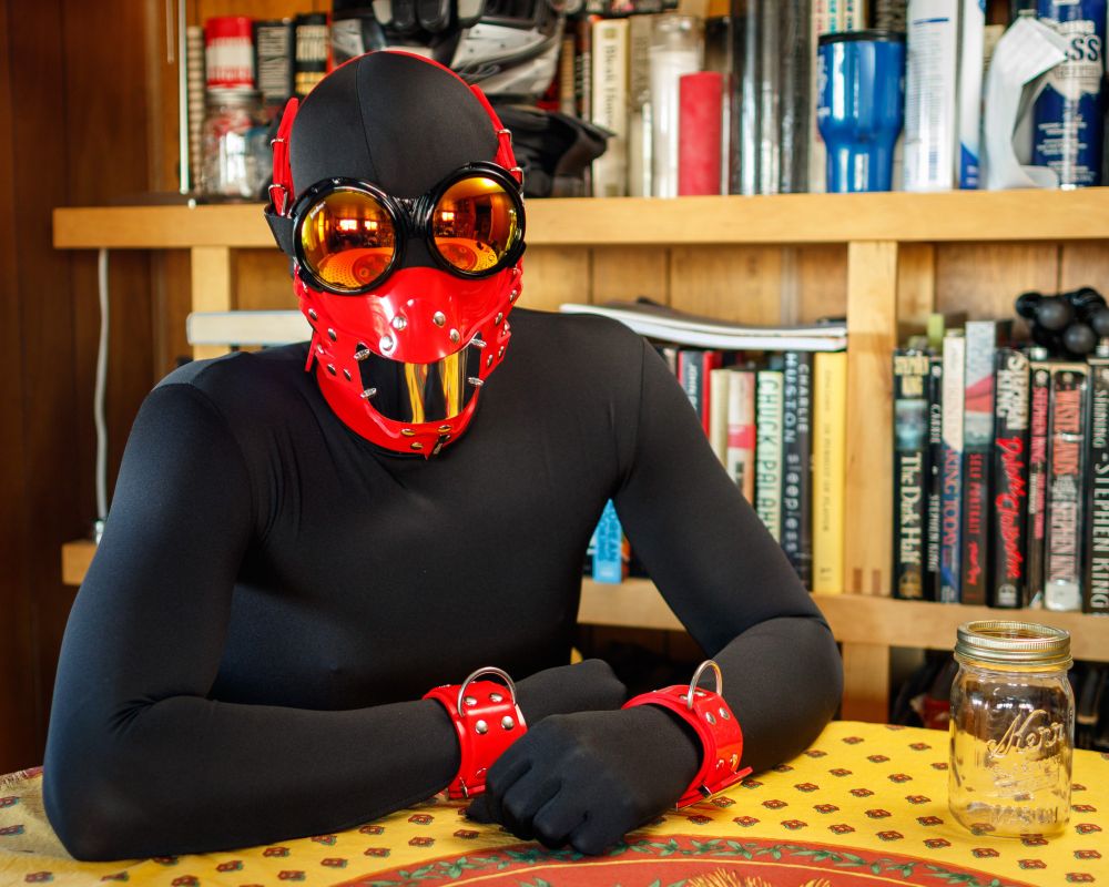 Wearing red silicone cuffs, collar, and muzzle over black spandex zentai.