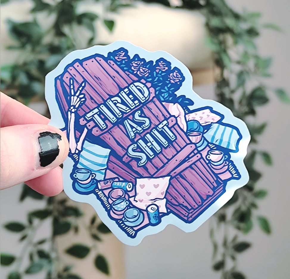 hand holding up a sticker of a wooden coffin titled "tired as shit"