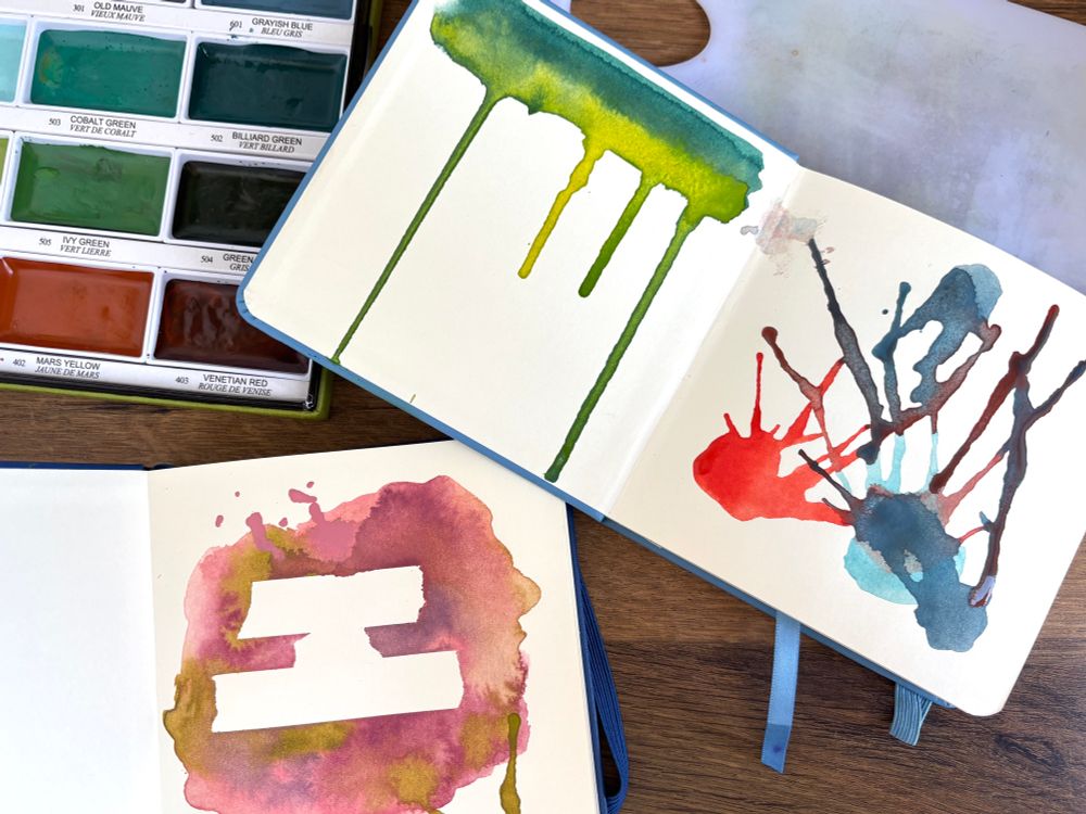 Two small sketchbooks on s as desk next to some paints. The sketchbook pages have very messy splashes and splotches of paint across them.