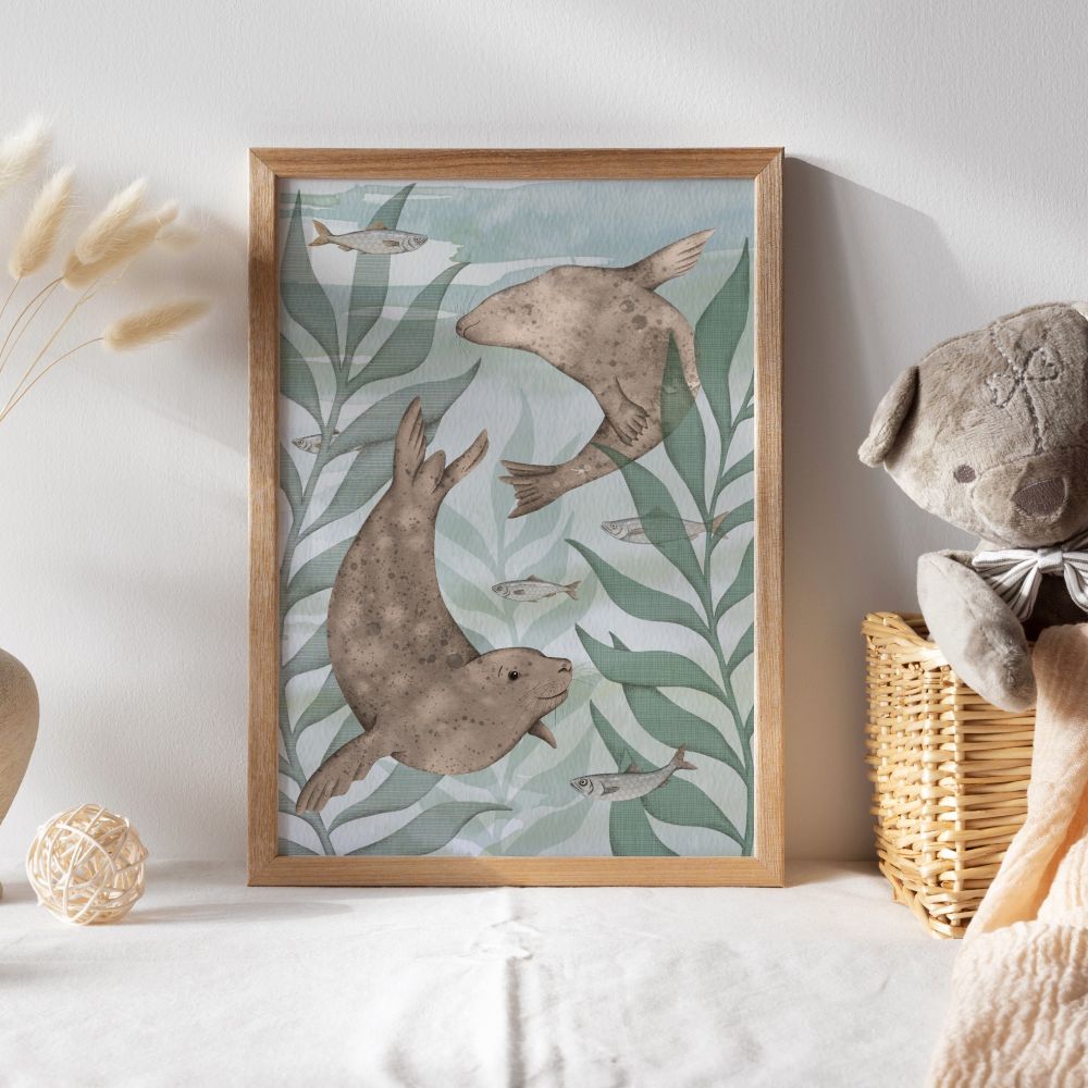 A wooden frame with an illustration in it of two seals swimming through green kelp fronds and turquoise water. There are fish amongst the kelp and next to the framed picture is a basket with a teddy bear in.