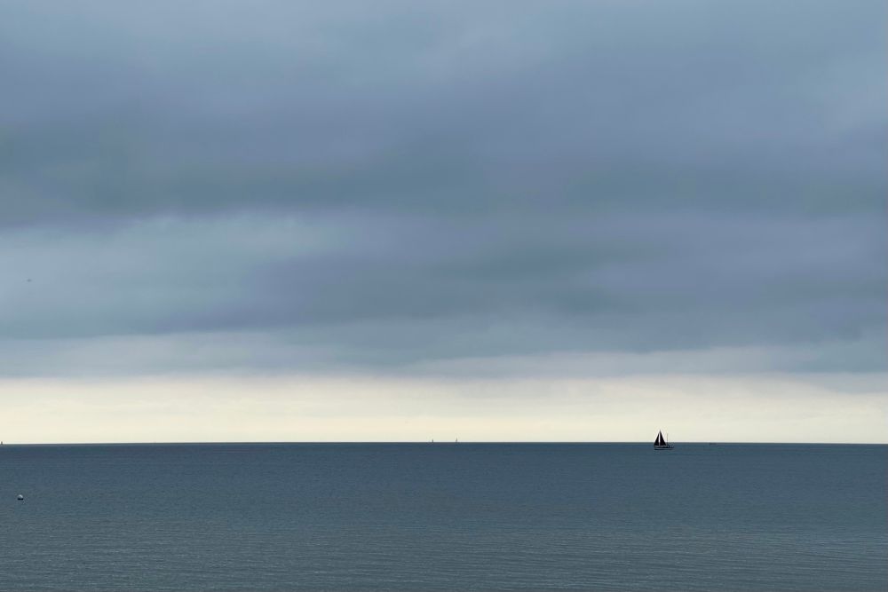 Farbfoto, Querformat: Panoramablick übers Meer, der Horizont teilt das Bild im unteren Viertel. Rechts im Bild ein kleines Segelboot im dunkelgrau schimmernden Wasser, darüber ein dunkelgrauer Himmel. Zwischen Wolkenbank und Horizont ein hellerer fast wolkenloser Streifen.