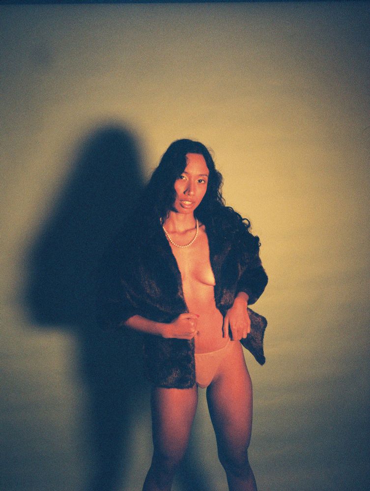 Kalham draped in a brown fur, tan bottoms, and a pearl necklace. She is standing.