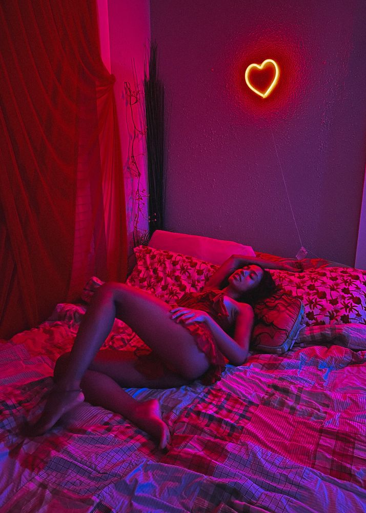 Kalham laying in bed in a red night gown illuminated by a red neon heart sign