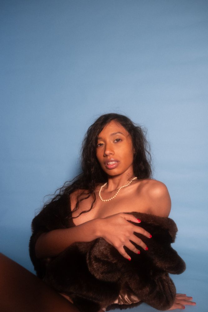 Portrait of Kalham against a blue background in a brown fur with a pearl necklace and her hair down. Her hand is held across her chest and the other is supporting her up.