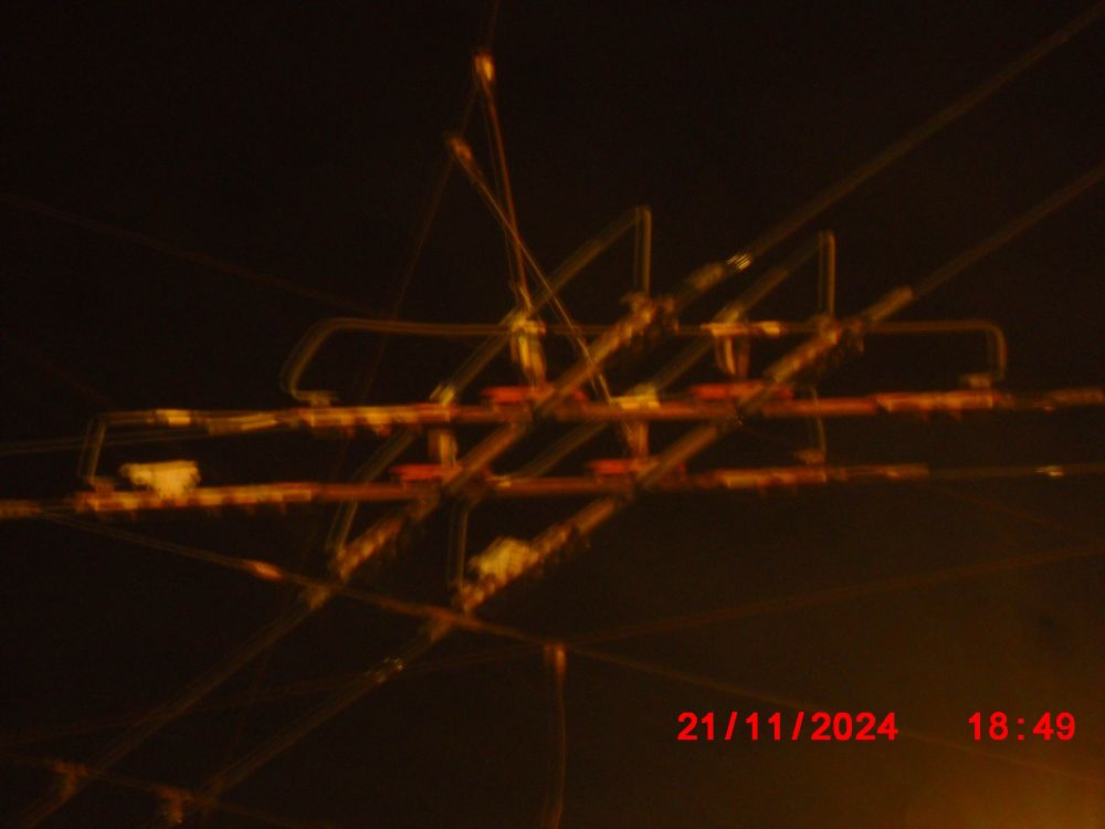 intersecting tram powerlines with orangey light illuminating the street somewhere out of frame. time is 18:49 and its vry dark