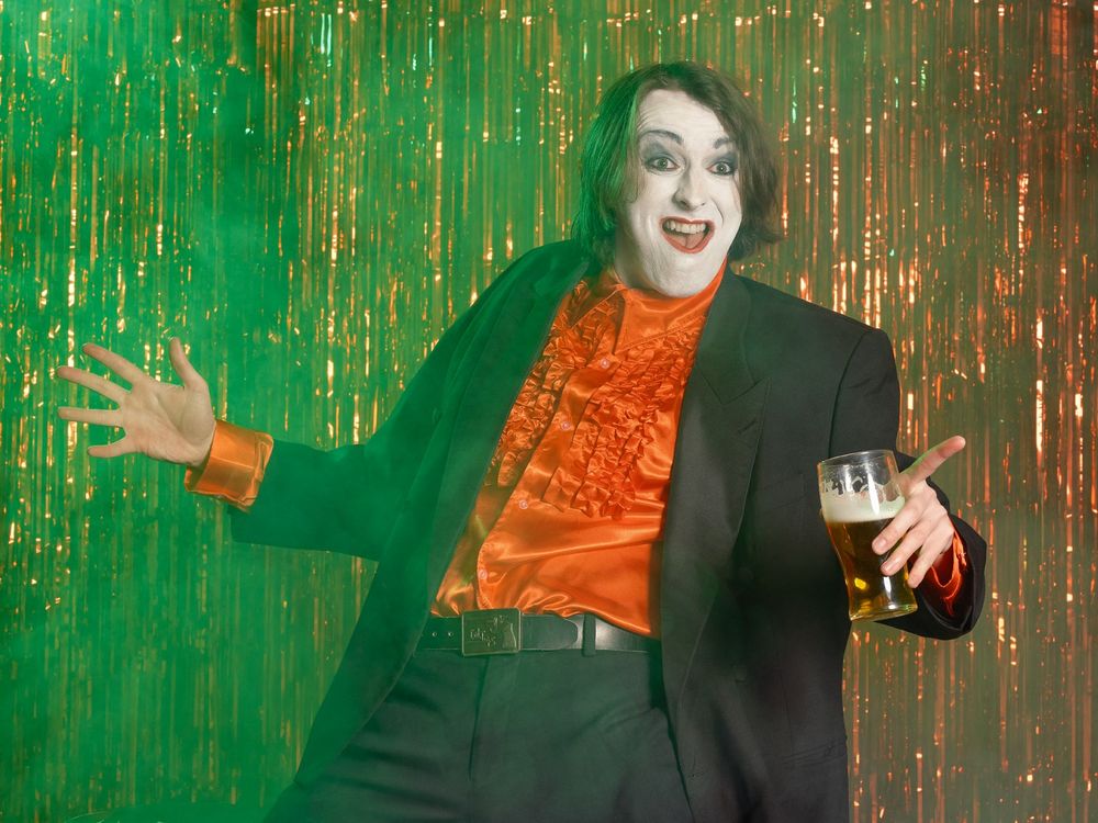 Production shot of Joe Kent-Walters as Frankie Monroe. Frankie has shoulder-length brown hair, his face is covered in white paint, with red lipstick and blue eye shadow. He is wearing a red, satin, ruffled shirt, black blazer and trousers. He is holding a pint glass of beer and looks like he is mid-show.