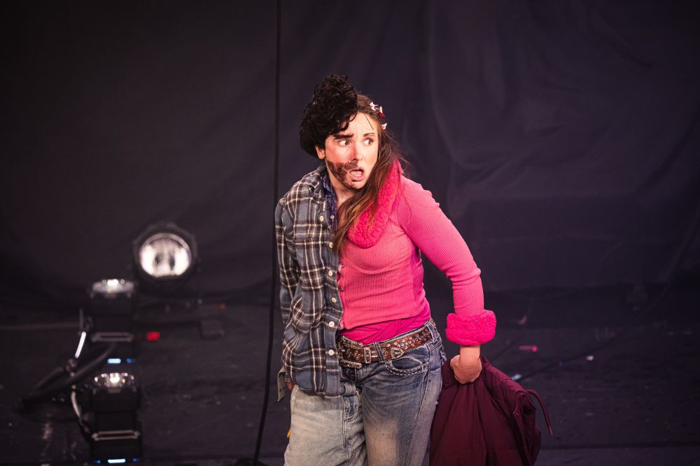 Production shot of Natalie Palamides' WEER. Natalie is split in half and playing two people. On her left side, she has short, dark, curly hair, a drawn on beard, a flannel shirt and jeans. On her right side, she has long, brown hair filled with butterfly clips. She is wearing a bright pink, furry, long-sleeve pink top and blue jeans.