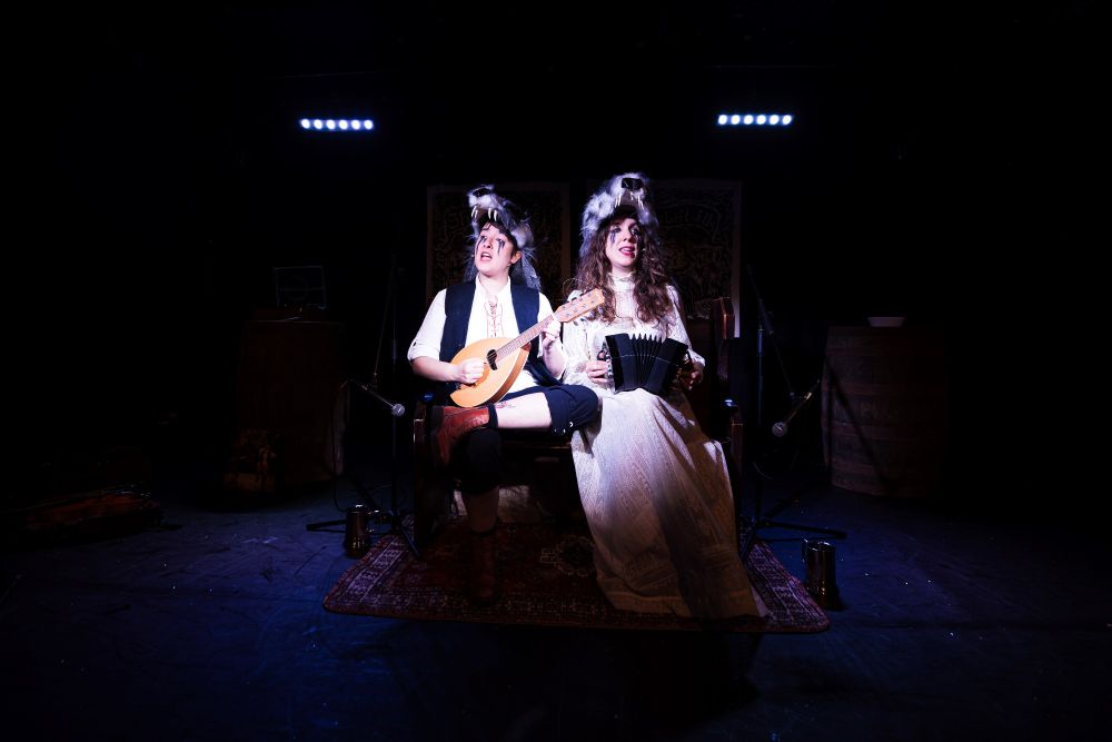 Production shot from Sh!t Theatre's Or What's Left Of Us. The cast of two are sat on stage on a bench, they are mid-song and have bagder's on their heads. They are playing a banjo and accordion. 