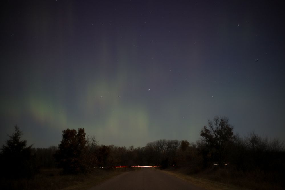 Aurora and stars appear in the sky above a narrow country road. The road ends at a nearby highway and lights from a passing vehicle on the highway are visible
