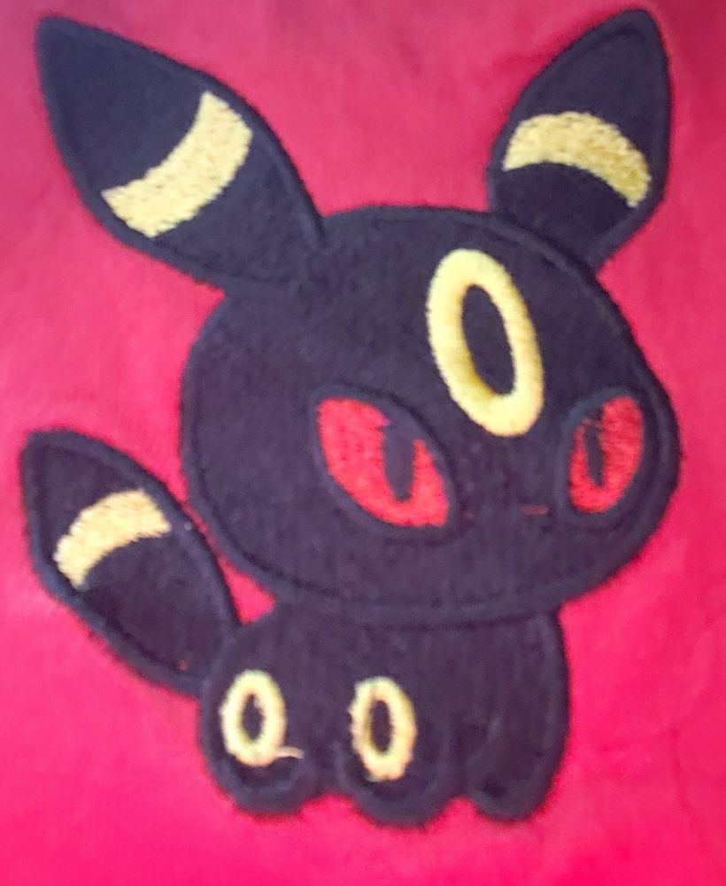 An image of a piece of embroidery on a red shirt of the pokemon Umbreon, a black fox like pokemon with yellow bands around its pointed ears and tail, yellow circles on its legs and forehead, and red eyes 