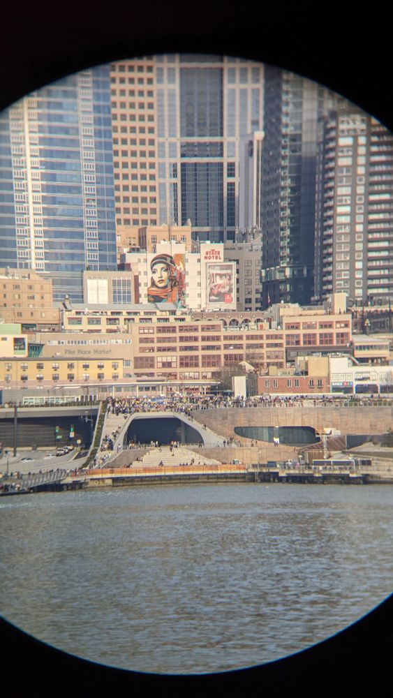 A photo of protesters standing along a pedestrian path and staircase overlooking the Seattle waterfront. Buildings tower over several hundred people who appear small as viewed from the ferry looking back at the city. A mural painting of a face on the State Hotel 🌹