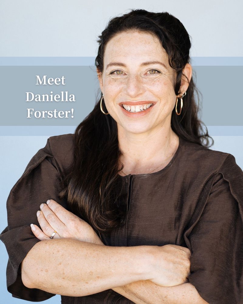 Dr Daniella Forster stands smiling with her arms crossed in front of a light blue background. She has long dark hair, gold hoop earrings, and is wearing a dark brown blouse. Overlaid text reads: “Meet Daniella Forster!”