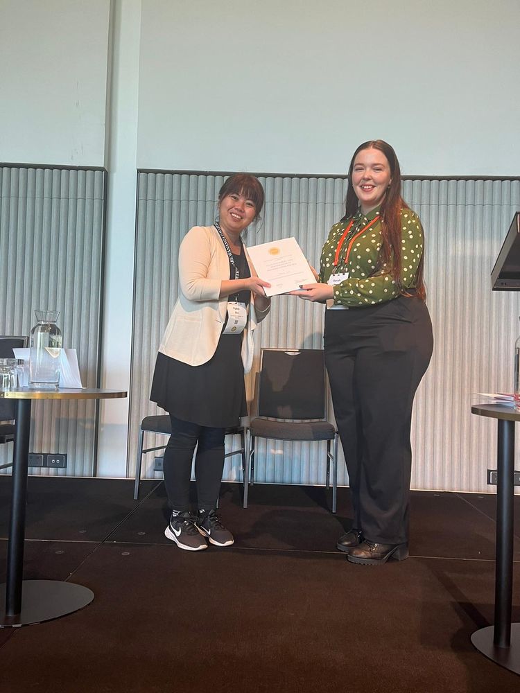 Two women on stage at the Christchurch Convention Centre. The woman on the left, wearing a light cardigan and sneakers, holds a certificate and smiles. The woman on the right, wearing a green polka-dot blouse, presents her with the PESA Doctoral and Postgraduate Presentation Award.