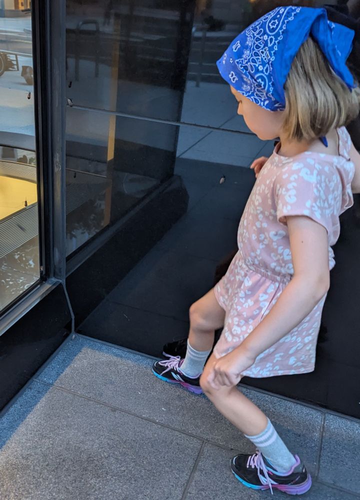 My child stomping spotted lantern flies on a sidewalk near a black reflective wall. There are several more lantern flies climbing up the wall