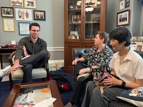 Three people talking in an office. A man is sitting in a chair, and two women are sitting on a couch facing him. One of the women has an adorable black Chihuahua in a pink harness on her lap, Evie. Evie is looking at the camera and is a very good dog.