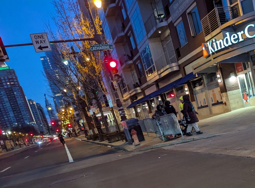Picture looking north on Broad St in early morning light with City Hall and green-lit skyscrapers in the background. There's a small group of fans standing behind a barricade on the sidewalk, some of them have folding chairs.
