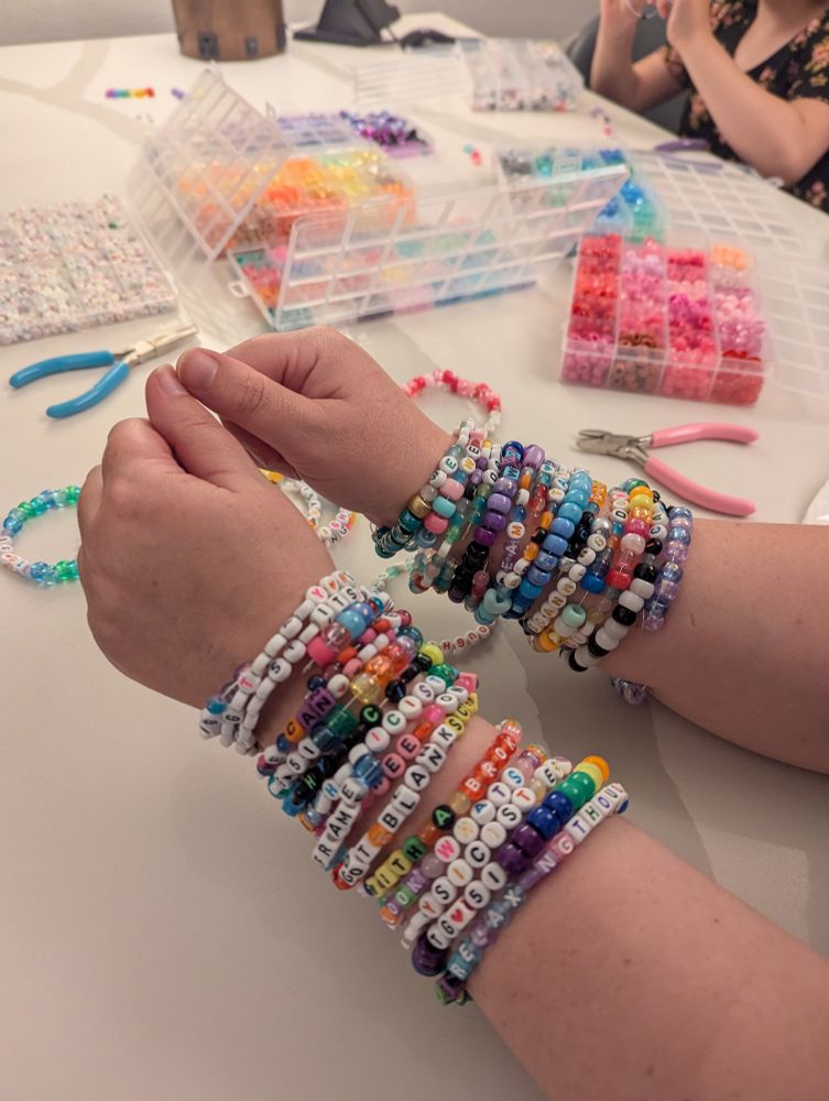 My arms, covered in beaded bracelets, leaning on a white table with more bead supplies in the background. The bracelets are Taylor Swift Eras Tour-inspired, with a Radiation Therapy twist. I'm so proud of my bracelet puns! Examples: Down Bad Crying At The Sim, I Can Do It With A Broken Cart, and TG <3 51