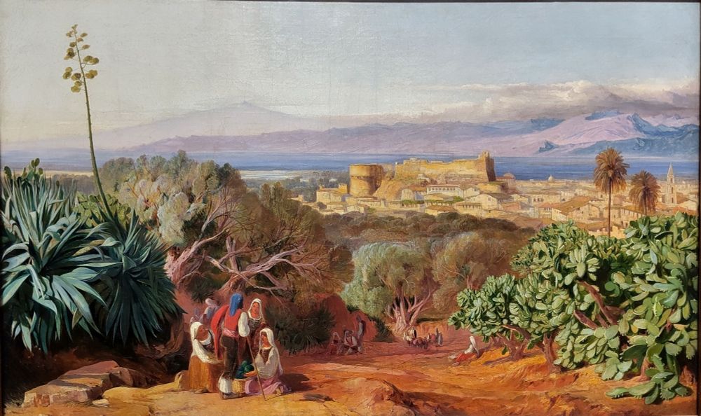 View from a wooded hilltop across a fortress above the city towards Sicily and quiet Mount Etna. In the foreground on the right an opuntia (disc cactus). On the left, a flowering aloe. To the right of the aloe a group of people; further down the slope towards the city more people.