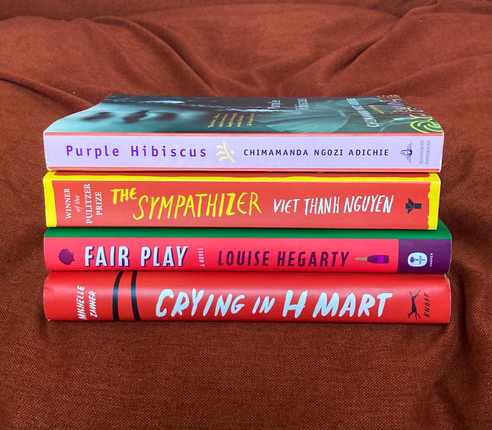 A stack of four books on an orange fabric chair: Crying in H Mart by Michelle Zavner, Fair Play by Louise Hegseth, The Sympathizer by Viet Thanh Nguyen, and Purple Hibiscus by Chimamanda Ngozi Adichie. 