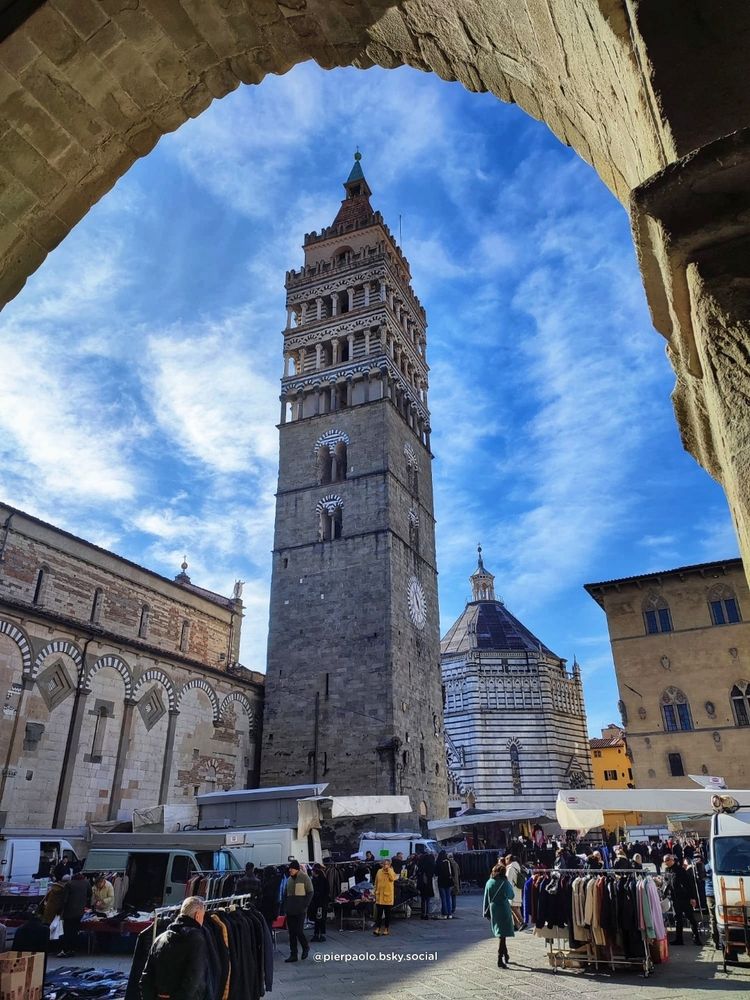 Foto della Piazza della Cattedrale a Pistoia (Toscana). Attraverso un arco del Palazzo Comune , nella piazza gremita di gente per il mercato cittadino, si vede la cattedrale il campanile, che si staglia al cento della foto, il battistero e parte del Palazzo Pretorio.