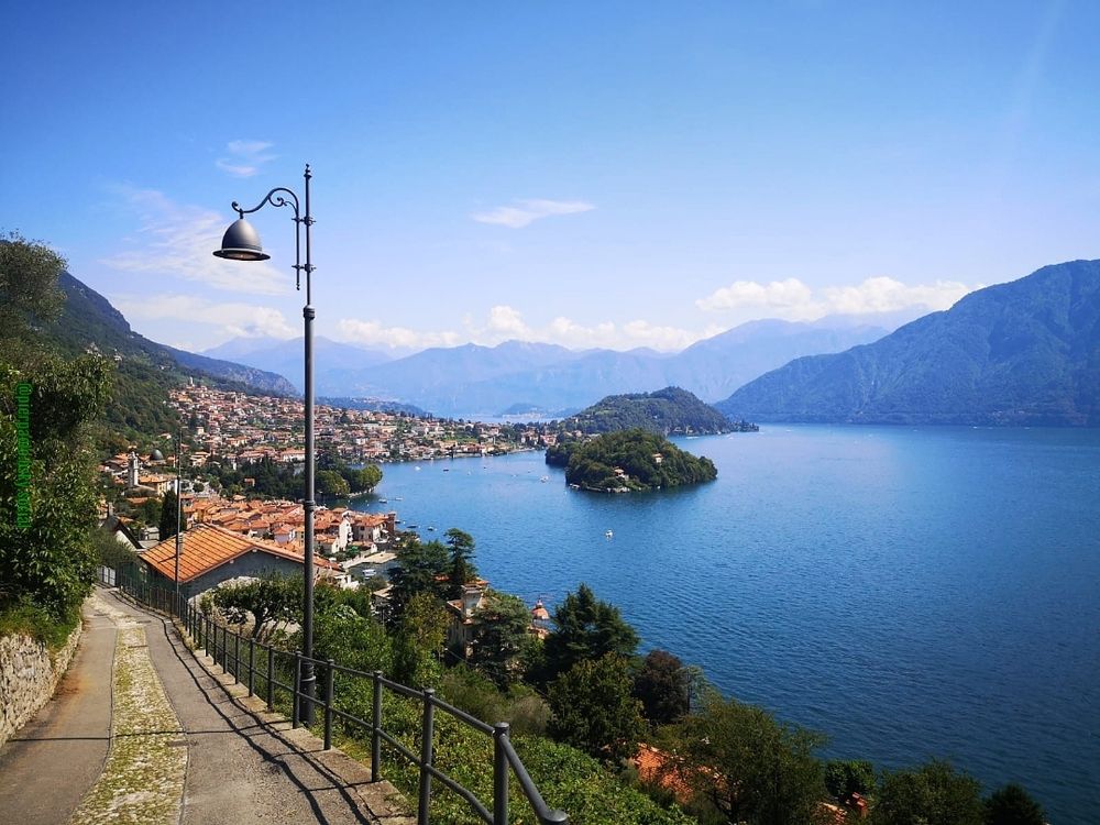 Dalla strada panoramica nei pressi di Colonno (Como), le case dei centri abitati di Lenno e Sala Comacina degradano dalle pendici dei monti fino alla riva del Lago di Como, nelle cui acque di un intenso azzurro, risalta il verde della vegetazione che ricopre l'Isola Comacina. Nel cielo azzurro, bianche nuvole  coronano le cime dei monti che circondano il lago.