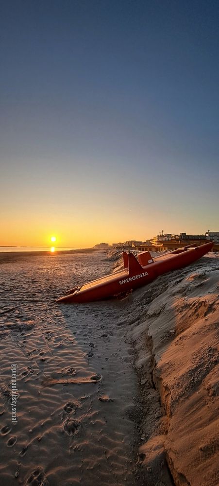 Un pattino di salvataggio di color rosso con la scritta emergenza appoggiato sul terrapieno di sabbia creato per evitare che le mareggiate possano erodere la spiaggia, è illuminato dai primi raggi del sole. La brina che ricopre la sabbia risplende al sole. In lontananza si vedono le sagome dei caseggiati dell'abitato di Valverde (Cesenatico).