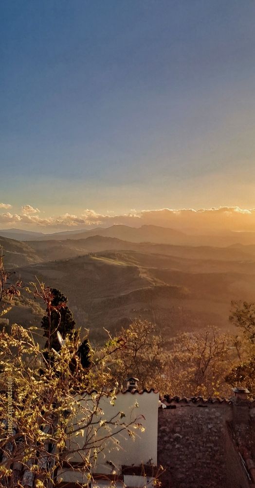 Oltre la chioma ormai spoglia di una robinia e i tetti di due abitazioni, le colline e le montagne, ricoperte da un leggero strato di foschia, sono illuminate dagli ultimi raggi dorati del sole al tramonto.